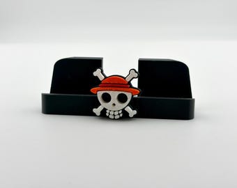 May include: A black plastic phone stand featuring a central emblem of a white skull with a red straw hat and crossed bones. The stand has a rectangular base with two upright supports, and the emblem is attached to the front center of the base.