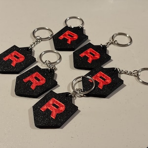 May include: A collection of black keychains with a red letter "R" on a shield-shaped design. Each keychain has a silver keyring attached. The keychains are arranged on a light-colored surface.