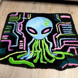 Puede incluir: Una alfombra rectangular negra con una cara de alienígena azul, ojos ovalados grandes y tentáculos verdes. El alienígena está rodeado por un diseño geométrico colorido en rosa, verde y amarillo.