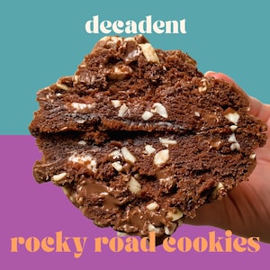 Puede incluir: Primer plano de una decadente galleta de rocky road, que muestra las chispas de chocolate, los malvaviscos y las nueces picadas. La galleta se sostiene en una mano, con la mitad inferior de la galleta rota, revelando el chocolate pegajoso en el interior.
