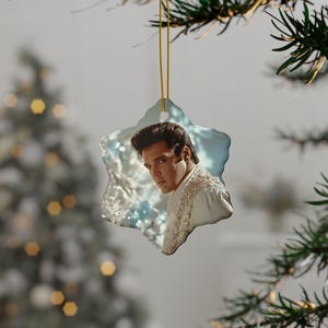 May include: A star-shaped Christmas ornament featuring a portrait of Elvis Presley. The ornament has a light blue and white frosted edge and a gold string for hanging. Elvis is wearing a white shirt with decorative embellishments.