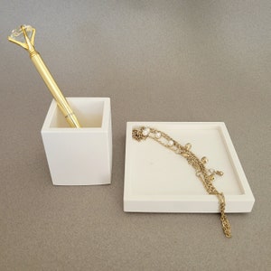 May include: Two white ceramic trays, one with a gold pen with a diamond-shaped top and the other with a gold chain necklace with pearl accents.