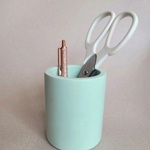 May include: A light green ceramic pencil holder with a pair of white scissors and a pink glitter pen inside.