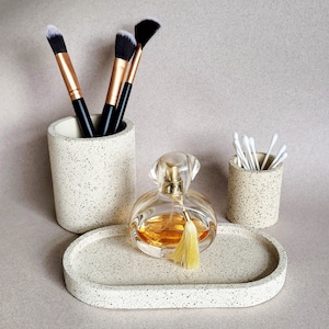 May include: A set of three speckled beige ceramic containers. The containers are a cylindrical makeup brush holder, a cylindrical cotton swab holder, and a rectangular tray. A clear glass bottle of perfume with a gold tassel sits on the tray.