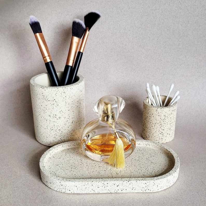 May include: A set of three speckled beige ceramic containers. The containers are a cylindrical makeup brush holder, a cylindrical cotton swab holder, and a rectangular tray. A clear glass bottle of perfume with a gold tassel sits on the tray.