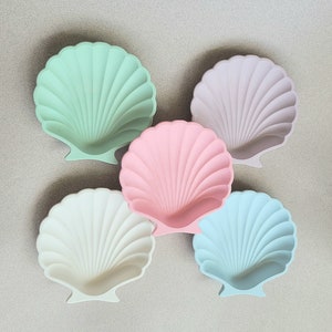 May include: Five seashell-shaped ceramic dishes in different colours. The dishes are light green, light purple, light pink, white, and light blue.