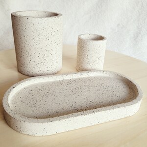 May include: A set of three speckled beige ceramic bathroom accessories: a tall cylindrical container, a shorter cylindrical container, and an oval tray.
