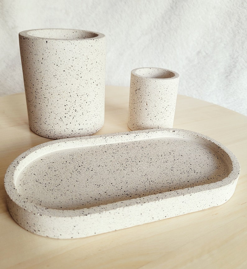 May include: A set of three speckled beige ceramic bathroom accessories: a tall cylindrical container, a shorter cylindrical container, and an oval tray.