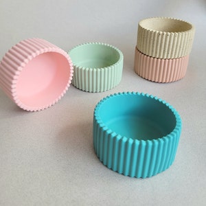 May include: A selection of small, round, ribbed concrete bowls in pastel shades of pink, mint green, light beige, and turquoise. The bowls feature a simple, modern design, suitable for holding small items or as decorative accents.