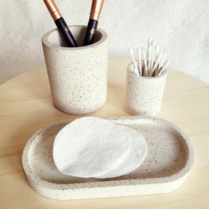 May include: A set of three speckled gray ceramic bathroom organizers. The set includes a round container for cotton swabs, a taller round container for makeup brushes, and a rectangular tray for cotton rounds.