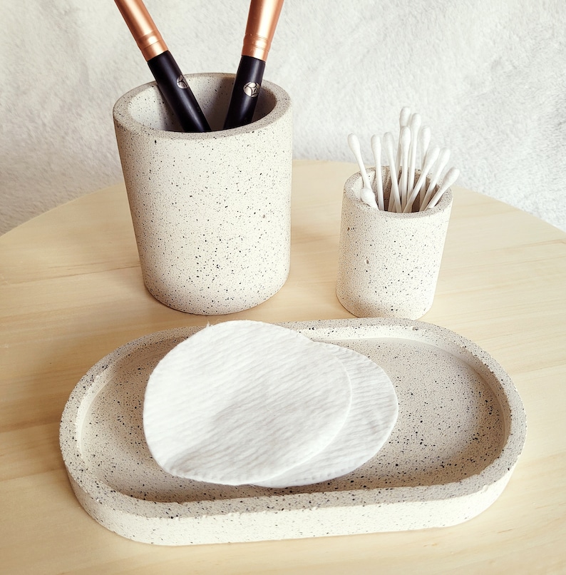 May include: A set of three speckled gray ceramic bathroom organizers. The set includes a round container for cotton swabs, a taller round container for makeup brushes, and a rectangular tray for cotton rounds.