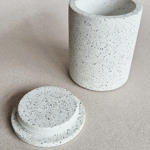 May include: A white speckled cylindrical container with a matching lid. The container has a slightly tapered shape and a wide opening. The lid is round and sits next to the container. The surface has a speckled pattern.