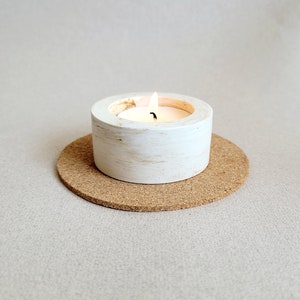 May include: A white candle holder with a distressed finish sits on a cork coaster. A small white tea light candle is burning inside the holder.