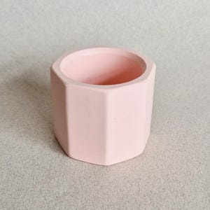 May include: A small, pale pink, geometric planter with a hexagonal design. The planter is made of a solid material and has a smooth, matte finish. The interior is the same colour as the exterior. Suitable for small succulents or other plants.