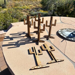 May include: A set of six wooden cactus-shaped ornaments. The ornaments are made of light brown wood and have a natural wood grain finish. The ornaments are arranged in a circle on a light brown surface.