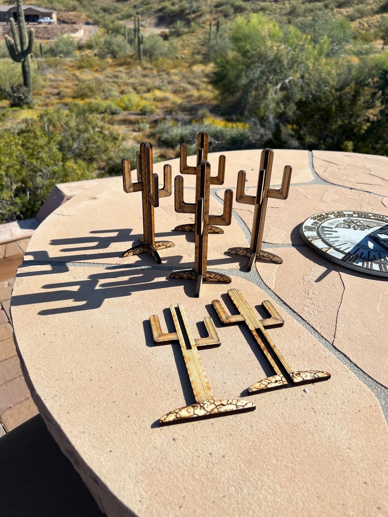 May include: A set of six wooden cactus-shaped ornaments. The ornaments are made of light brown wood and have a natural wood grain finish. The ornaments are arranged in a circle on a light brown surface.