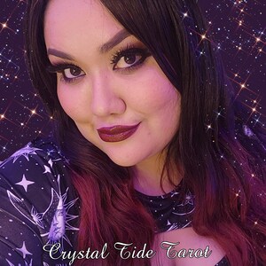May include: A woman with long dark hair and burgundy highlights is wearing a black and white patterned shirt with a crescent moon and stars. The text "Crystal Tide Tarot" is written in a cursive font at the bottom of the image.