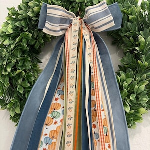 May include: A decorative bow featuring blue and white striped fabric, layered with long ribbons in solid blue, solid olive green, orange gingham, and a cream-colored ribbon printed with small orange pumpkins and blue leaves, attached to a green wreath.
