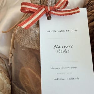 May include: A burlap drawstring pouch containing dried fruit slices, tied with a striped orange and white ribbon. A white rectangular tag attached to the ribbon reads Allyn Lane Studio, Harvest Cider, Aromatic Stovetop Simmer, Simmer Pot Blend, Handcrafted, Small Batch.