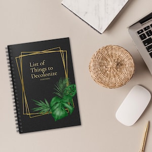 May include: A black spiral bound notebook with a gold frame and the text "List of Things to Decolonize EVERYTHING" on a white background with a laptop, a mouse, a coaster, and a pen.