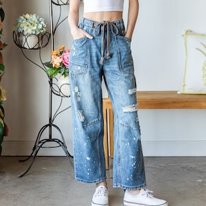 Peut inclure: Une femme porte un haut court blanc et un jean bleu large d&eacute;lav&eacute; avec des &eacute;claboussures de peinture. Elle porte des baskets blanches. Un support &agrave; plantes en m&eacute;tal noir avec des fleurs est en arri&egrave;re-plan.