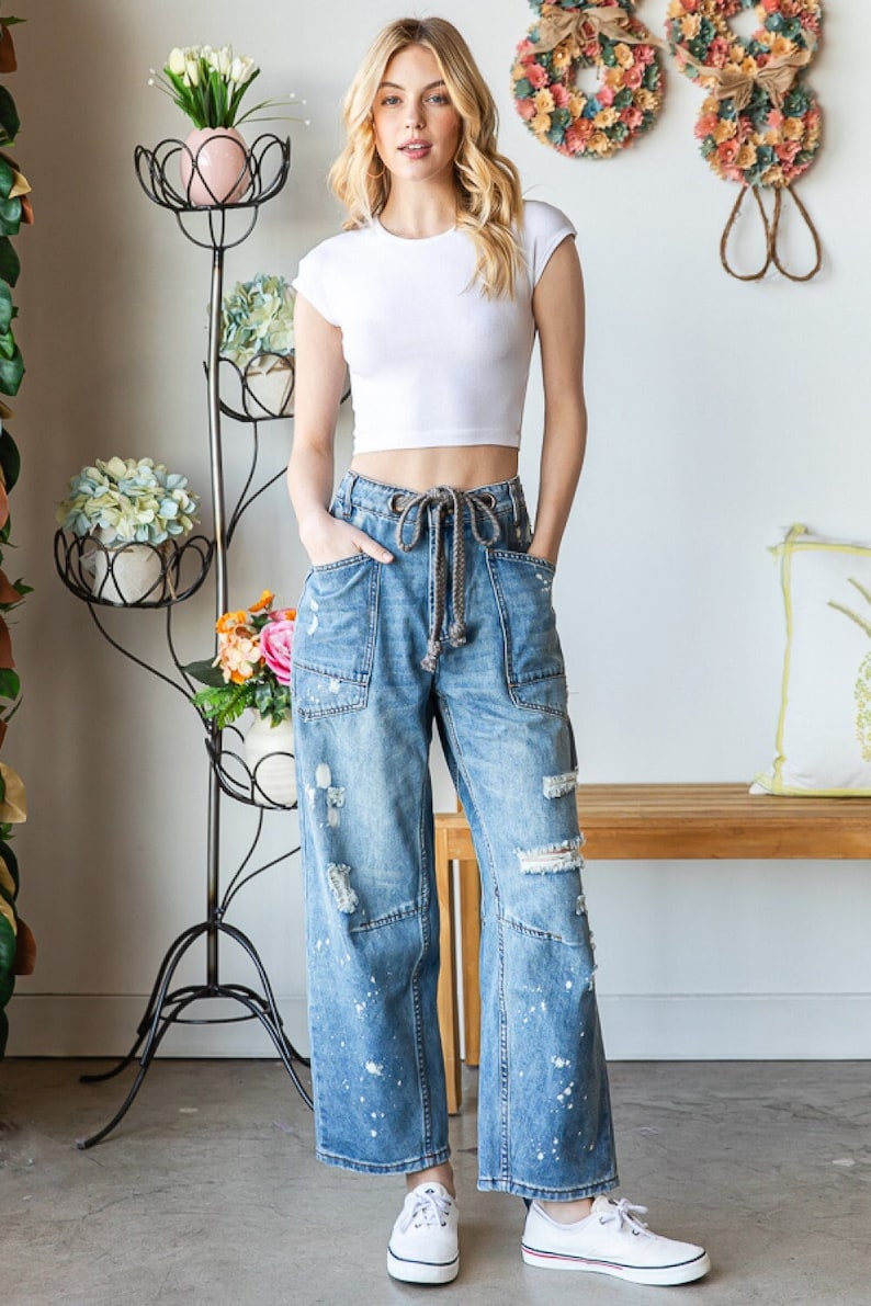 Peut inclure: Une femme porte un haut court blanc et un jean bleu large d&eacute;lav&eacute; avec des &eacute;claboussures de peinture. Elle porte des baskets blanches. Un support &agrave; plantes en m&eacute;tal noir avec des fleurs est en arri&egrave;re-plan.