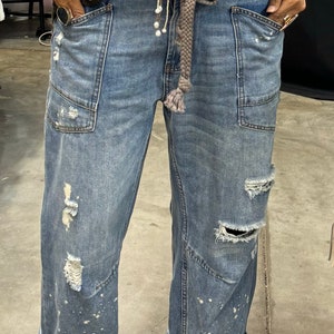 Peut inclure: Jean bleu clair avec des d&eacute;tails us&eacute;s, dont des d&eacute;chirures et des &eacute;claboussures de peinture. Le jean a de grandes poches avant et est retrouss&eacute; aux chevilles. Une ceinture tress&eacute;e et un pull vert fonc&eacute; compl&egrave;tent le look. Des sandales marron sont port&eacute;es.