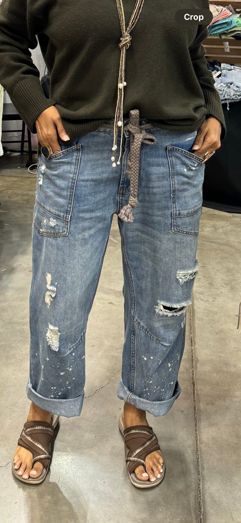 Peut inclure: Jean bleu clair avec des d&eacute;tails us&eacute;s, dont des d&eacute;chirures et des &eacute;claboussures de peinture. Le jean a de grandes poches avant et est retrouss&eacute; aux chevilles. Une ceinture tress&eacute;e et un pull vert fonc&eacute; compl&egrave;tent le look. Des sandales marron sont port&eacute;es.