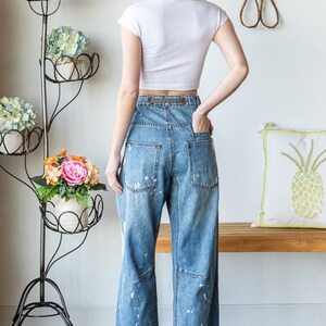 Peut inclure: Une femme porte un haut court blanc et un jean bleu clair &agrave; jambes larges avec des &eacute;claboussures de peinture blanche. Le jean a une taille haute et une boucle de ceinture d&eacute;corative. Des baskets blanches compl&egrave;tent la tenue. Un porte-plantes en m&eacute;tal noir avec des fleurs est en arri&egrave;re-plan.