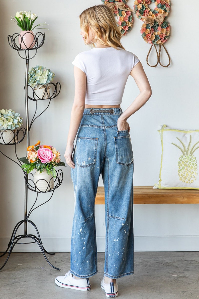 Peut inclure: Une femme porte un haut court blanc et un jean bleu clair &agrave; jambes larges avec des &eacute;claboussures de peinture blanche. Le jean a une taille haute et une boucle de ceinture d&eacute;corative. Des baskets blanches compl&egrave;tent la tenue. Un porte-plantes en m&eacute;tal noir avec des fleurs est en arri&egrave;re-plan.