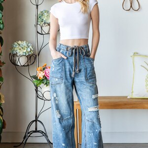 Peut inclure: Une femme porte un haut court blanc et un jean bleu large et d&eacute;lav&eacute; avec une taille &agrave; cordon. Le jean pr&eacute;sente des patchs et des &eacute;claboussures de peinture blanche. Des baskets blanches compl&egrave;tent la tenue. Un support &agrave; plantes en m&eacute;tal noir avec des fleurs est en arri&egrave;re-plan.