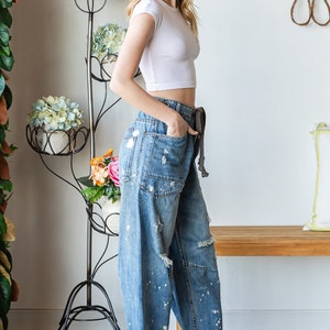 Peut inclure: Une femme porte un crop top blanc et un jean en denim d&eacute;lav&eacute; clair avec des &eacute;claboussures de peinture et des d&eacute;tails d&eacute;chir&eacute;s. Le jean a une taille &agrave; cordon et est associ&eacute; &agrave; des baskets blanches. Un support de plante en m&eacute;tal noir avec des fleurs est en arri&egrave;re-plan.