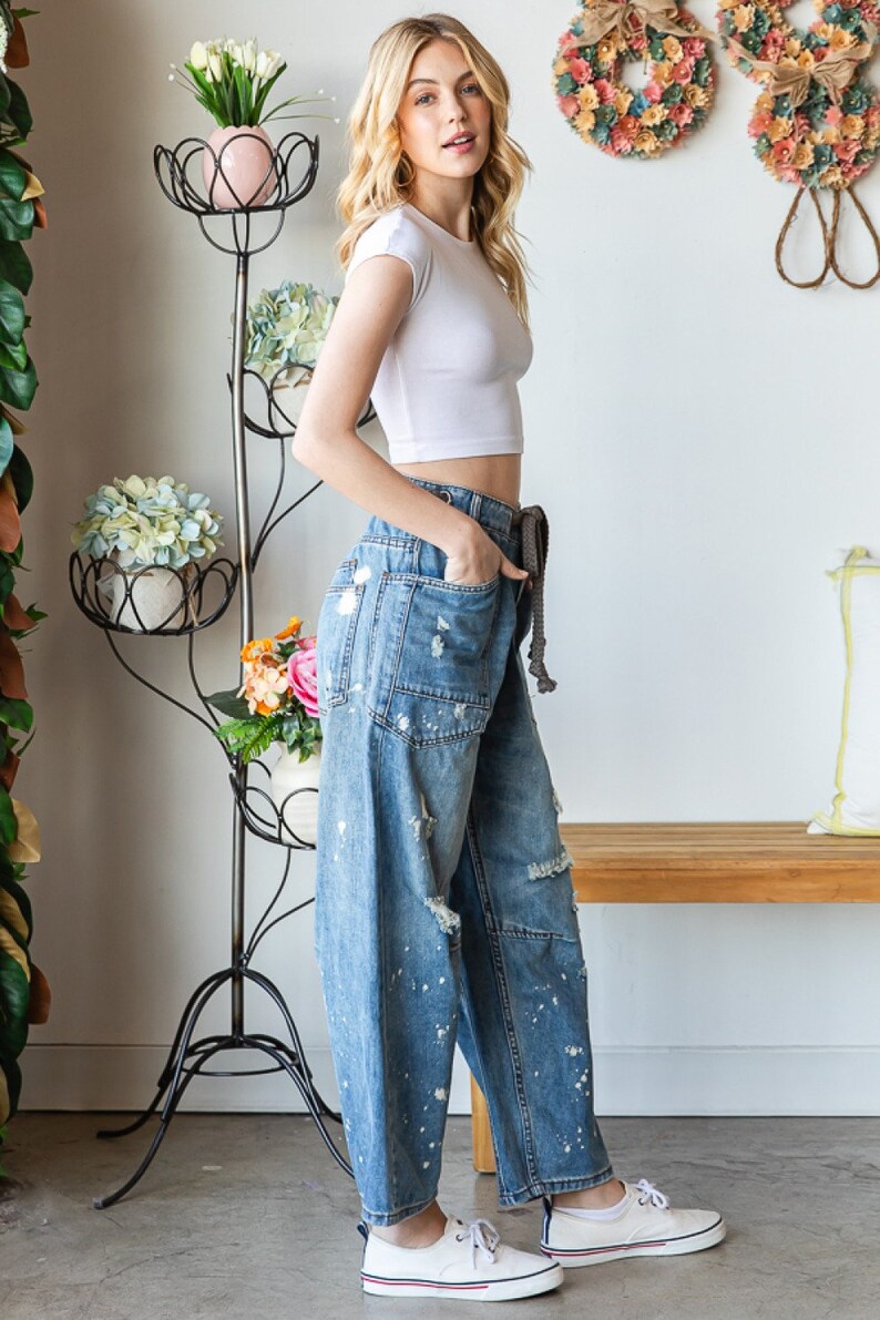 Peut inclure: Une femme porte un crop top blanc et un jean en denim d&eacute;lav&eacute; clair avec des &eacute;claboussures de peinture et des d&eacute;tails d&eacute;chir&eacute;s. Le jean a une taille &agrave; cordon et est associ&eacute; &agrave; des baskets blanches. Un support de plante en m&eacute;tal noir avec des fleurs est en arri&egrave;re-plan.
