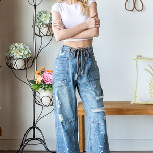 Peut inclure: Une femme porte un haut court blanc et un jean en denim bleu clair avec des d&eacute;tails d&eacute;chir&eacute;s et des &eacute;claboussures de peinture blanche. Le jean a une taille &agrave; cordon. Elle porte des baskets blanches. Un support &agrave; plantes en m&eacute;tal noir avec des fleurs est en arri&egrave;re-plan.