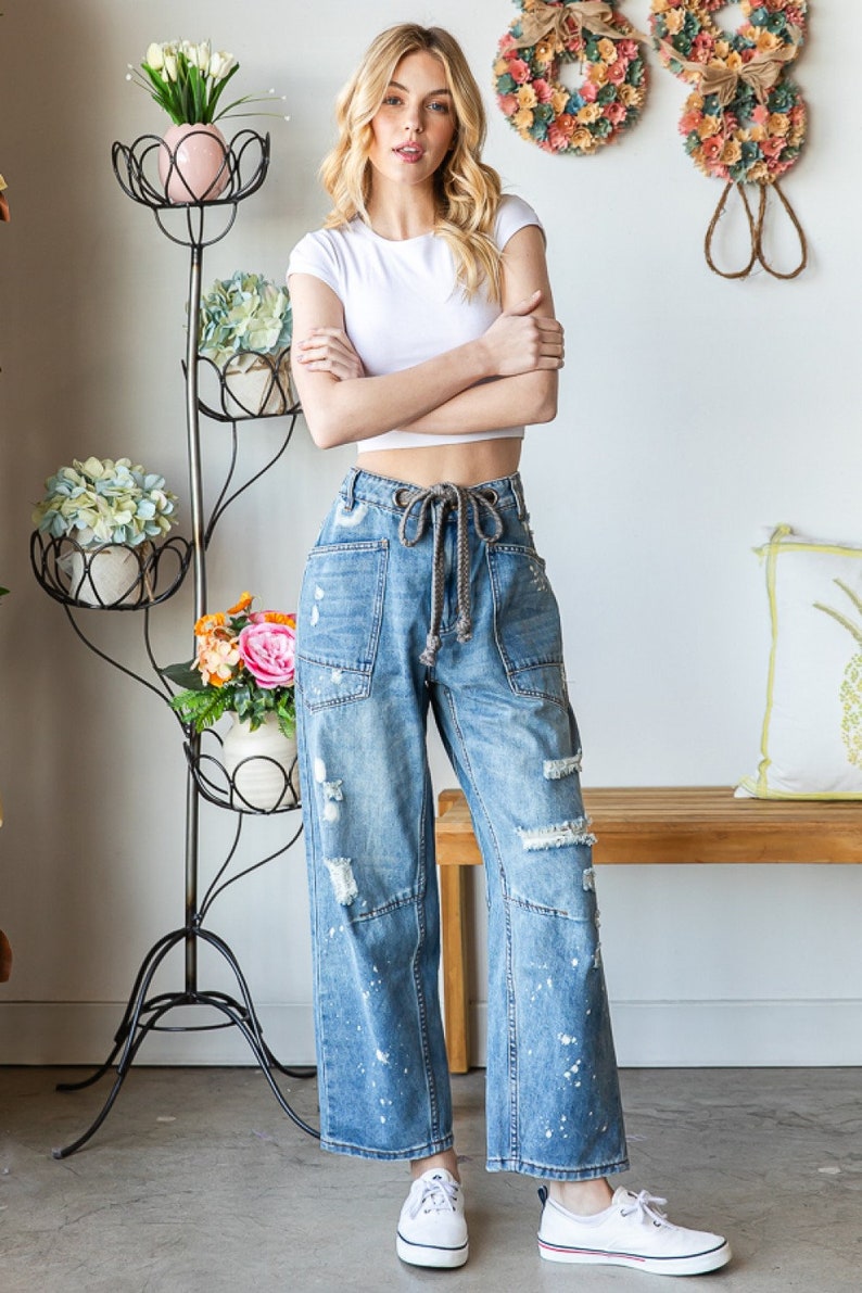 Peut inclure: Une femme porte un haut court blanc et un jean en denim bleu clair avec des d&eacute;tails d&eacute;chir&eacute;s et des &eacute;claboussures de peinture blanche. Le jean a une taille &agrave; cordon. Elle porte des baskets blanches. Un support &agrave; plantes en m&eacute;tal noir avec des fleurs est en arri&egrave;re-plan.