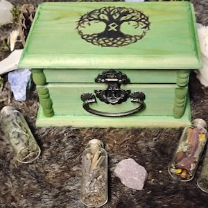 May include: A light green wooden box with a dark brown tree of life symbol on the lid and a metal latch. Three small clear glass vials containing dried herbs and botanical materials are positioned in front of the box on a dark textured surface.