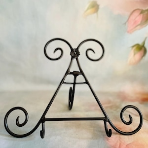 May include: A dark brown metal easel with a triangular frame and decorative scrollwork. The easel features a small flower detail at the top and curved hooks for holding items. The base has decorative swirls on each side.