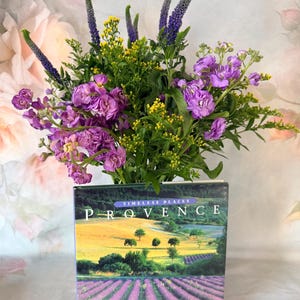 May include: A hardcover book titled Timeless Places Provence by Alexandra Bonfante-Warren, featuring a landscape cover of lavender fields, topped with a vibrant floral arrangement of purple stock flowers, yellow solidago, and purple veronica.
