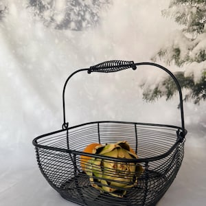 May include: A black wire basket with a rectangular base and rounded corners, featuring a single arched handle with a coiled metal grip. Inside the basket sits a single green artichoke and a round orange fruit.