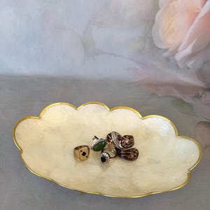 May include: An oval, scalloped-edge trinket dish with a pearlescent, off-white finish and a thin gold-toned rim. Resting in the center are several rings, including one with a black spade symbol, a green stone, and a dark, patterned band.