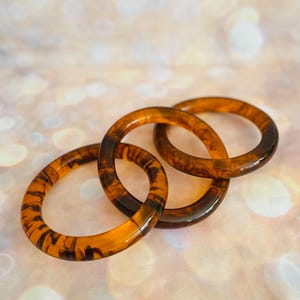 May include: Three amber-colored bangles with a marbled pattern. The bangles are made of a translucent material, with dark brown streaks throughout. The bangles are stacked together, creating a layered effect. The background is a soft, blurred bokeh of warm colors.
