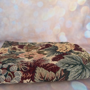 May include: A folded tapestry with a floral pattern in shades of burgundy, beige, green, and pink. The fabric features a detailed woven design with leaves and flowers, creating a vintage aesthetic. The background is a soft, blurred bokeh.
