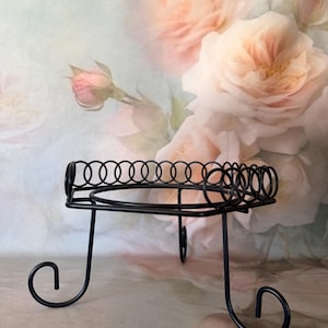 May include: A black metal stand featuring a square top with a decorative circular wire border and three legs that terminate in curled feet. The structure includes a central circular ring support designed to hold items securely in place.
