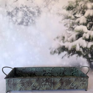 May include: A rectangular metal tray with a dark green and bronze patina finish, featuring an embossed botanical pattern of leaves and flowers. The tray has two curved side handles and stands on four small, rounded metal feet.
