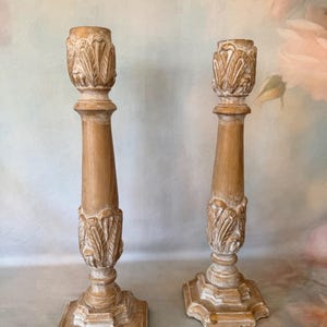 May include: Two tall wooden candle holders with a light brown finish and white distressed detailing. Each holder features a square base, a slender central column, and carved leaf-like patterns at the top and bottom sections of the stem.