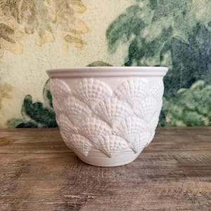 May include: A white ceramic planter with a seashell design. The planter has a rounded shape and a slightly flared rim. The seashell pattern covers the entire exterior of the planter, creating a textured surface. The planter is sitting on a wooden surface.
