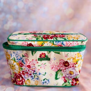 May include: A floral print cosmetic bag with a matching lid. The bag features a cream-colored background with a vibrant pattern of pink, red, yellow, and blue flowers. The bag has a green zipper and trim. The word "Celebrity" is printed on the bag.