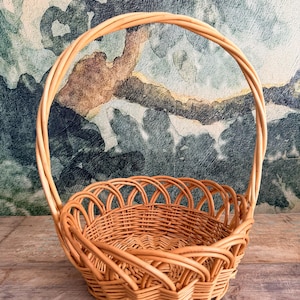 May include: A light brown wicker basket with a handle. The basket has a round base and a woven design. The handle arches over the top of the basket. The basket is sitting on a wooden surface.