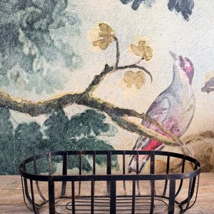 May include: A black metal basket with an oval shape and vertical bars. The basket sits on a wooden surface, with a painted mural of a bird and floral branches in the background. The basket is empty.