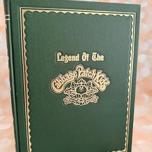 May include: A hardcover book with a dark green cover and gold embossed lettering. The title "Legend Of The Cabbage Patch Kids" is displayed in gold. The cover has a gold border.